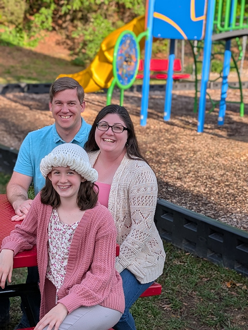 Shannon with her husband and daughter seated at a picnic table with colorful playground equipment in the background